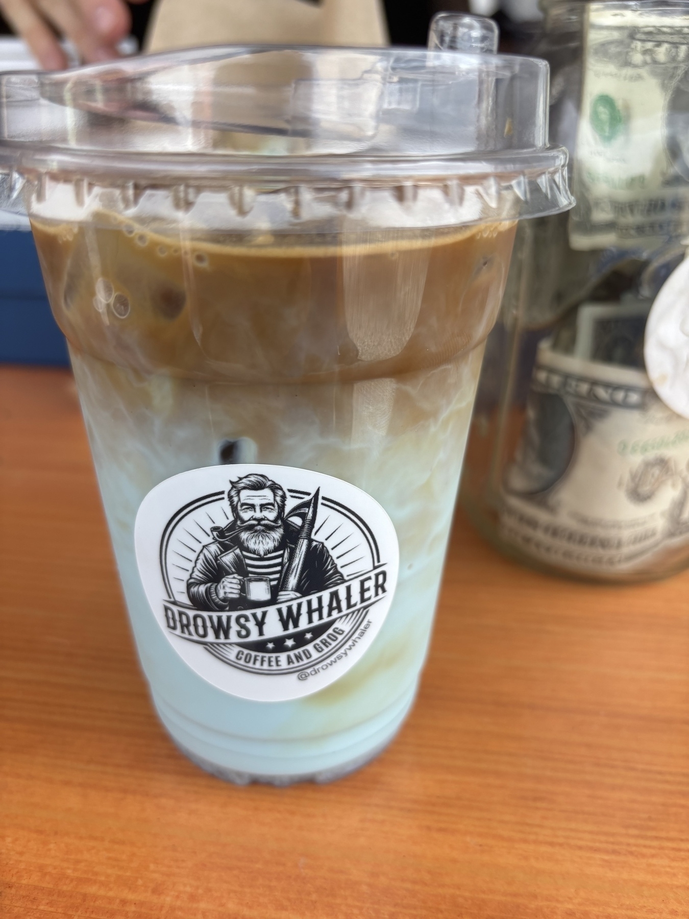 A blue milk iced espresso in a clear plastic take-out glass. The espressso sits on top of the blue milk. There is a “Drowsy Whaler” sticker on the glass and a tip jar to the right. 