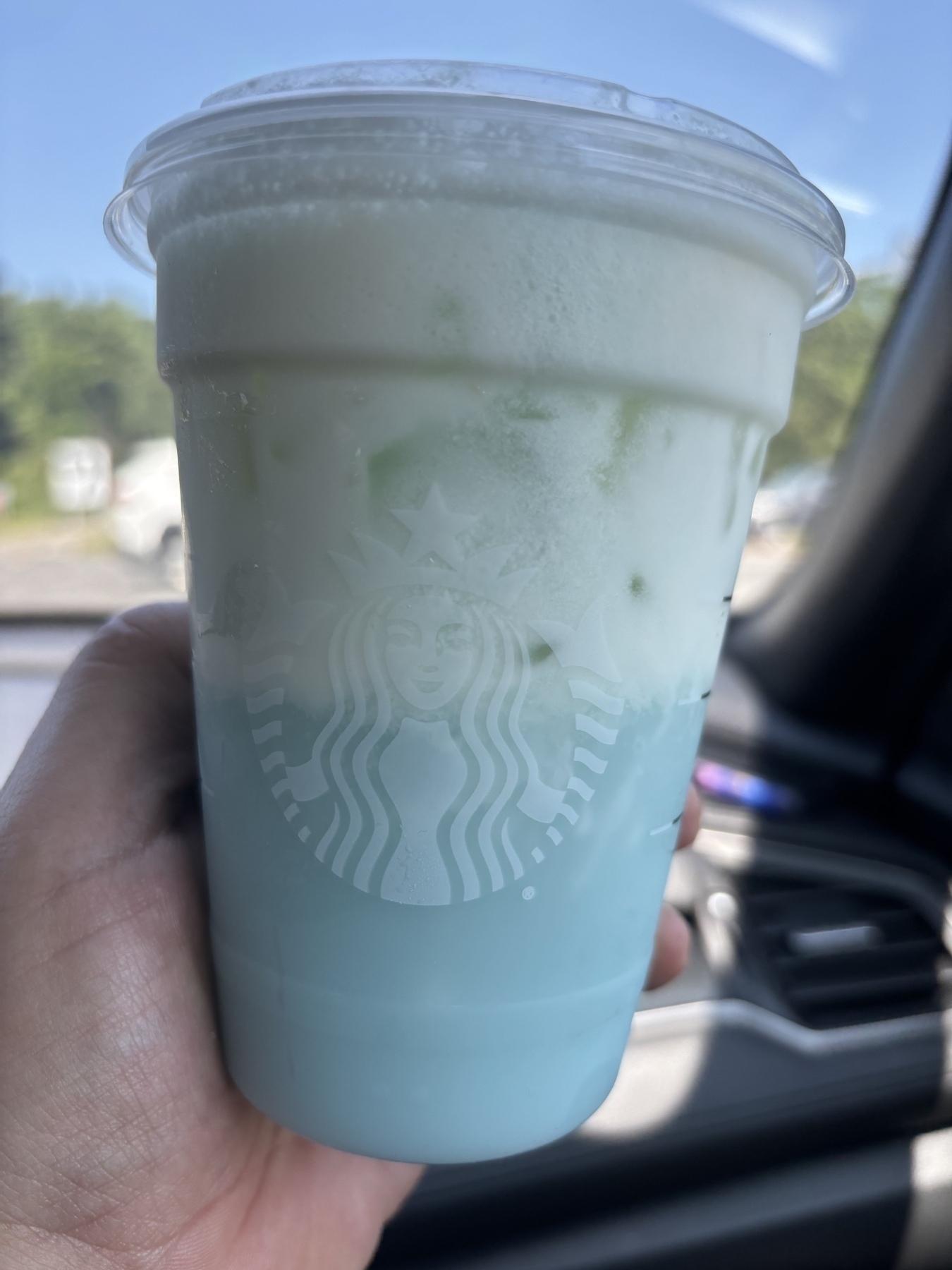A Starbucks drink held up in a car looking out the window. The drink is blue with a white top, making it look like a gummy shark