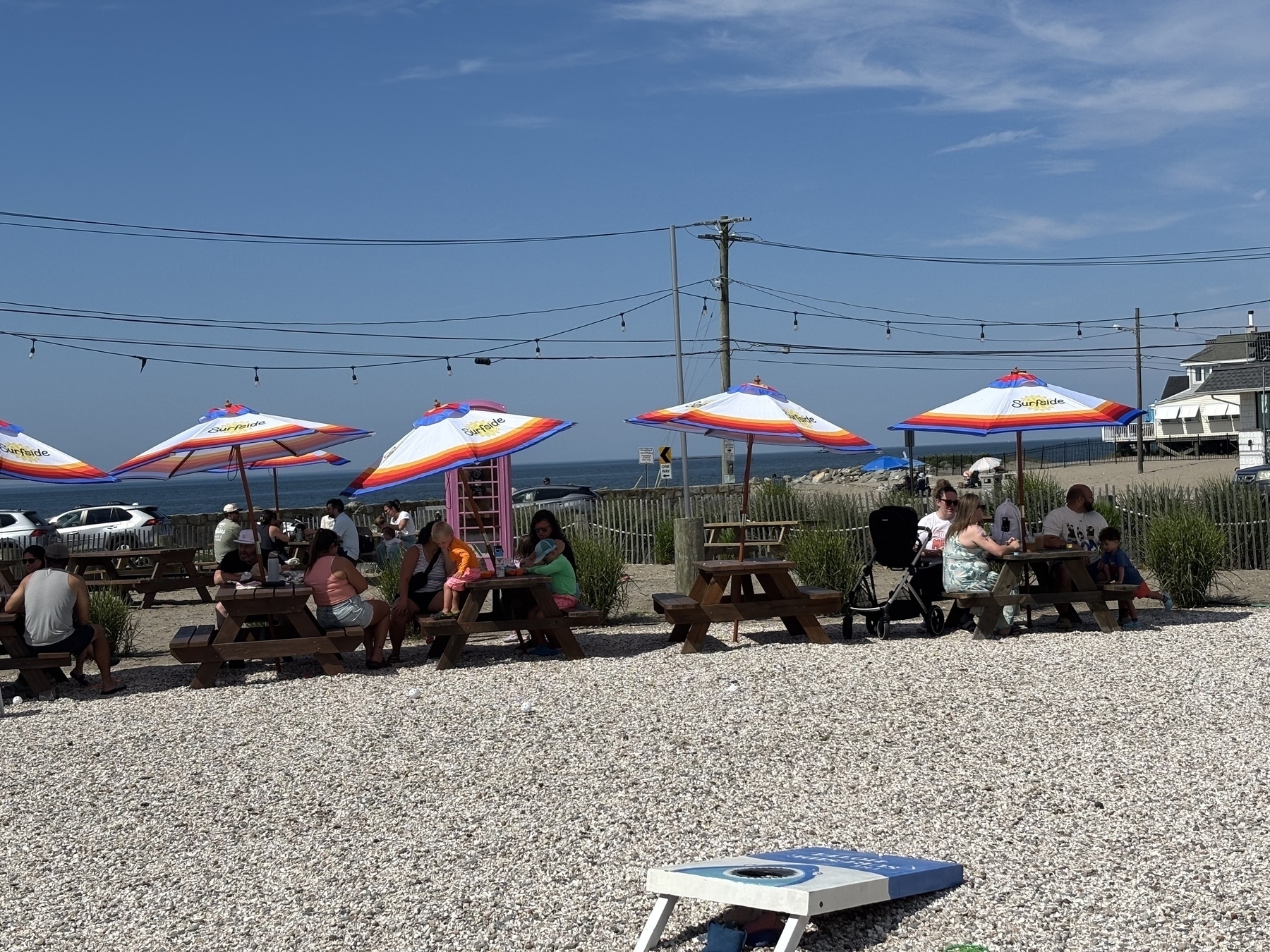 A line of picnic tables with colorful umbrellas on a sandy beach area. A Seawall and ocean with blue skies are in the background. 