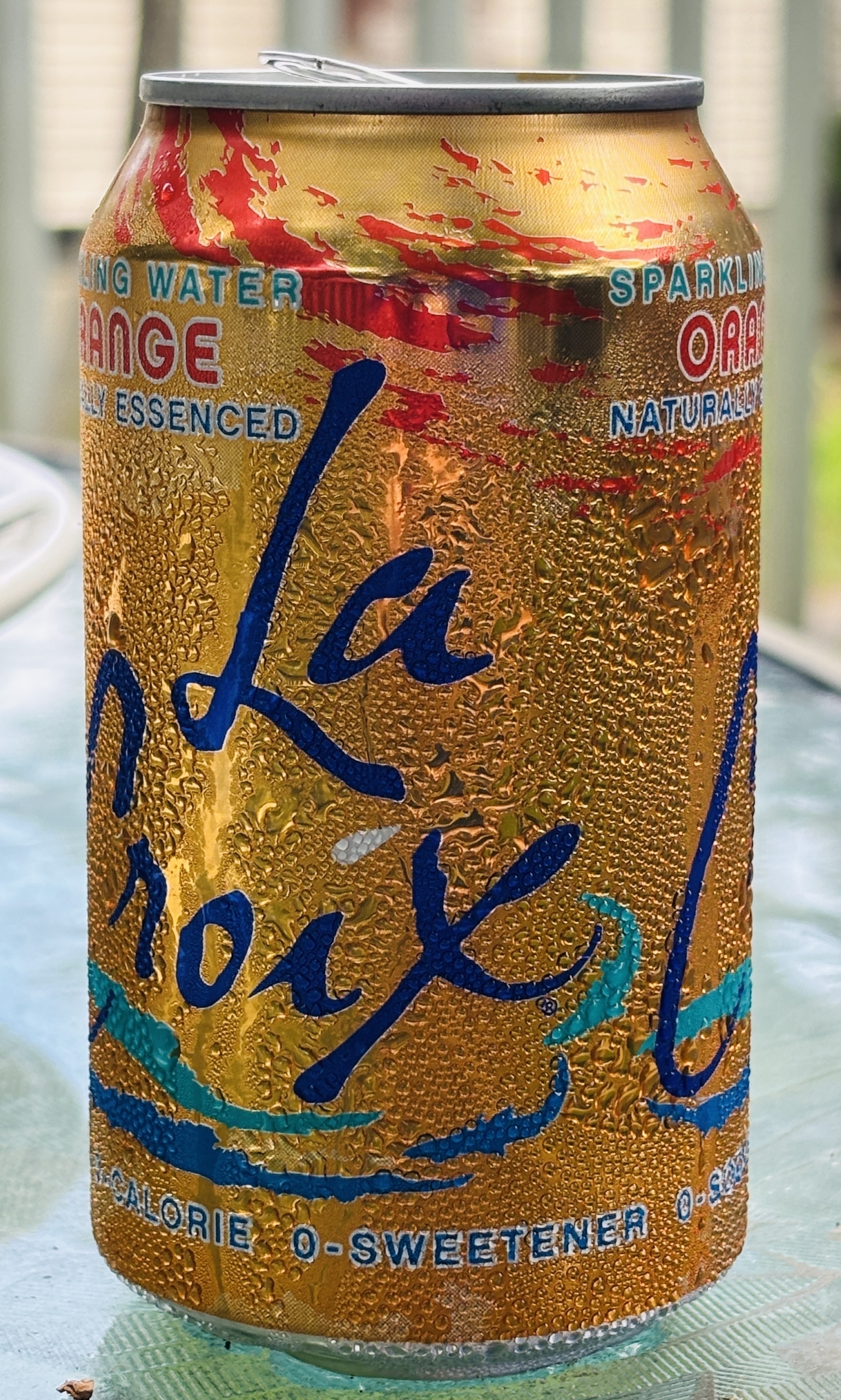 An orange can of La Croix orange seltzer sits on a table on a deck. Beads of condensation cover the can, giving it a cool enticing vibe. 