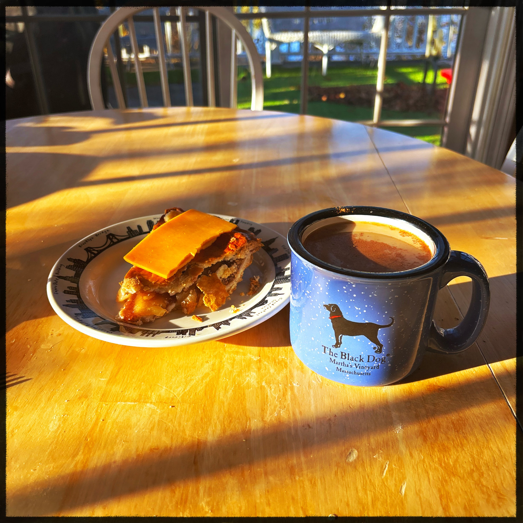 A slice of apple pie with a slice of cheddar cheese sits atop of a diner plate. Next to it is a cup of coffee in a blue Black Dog mug. Sunlight streams across the table from the left giving a warm glow. 