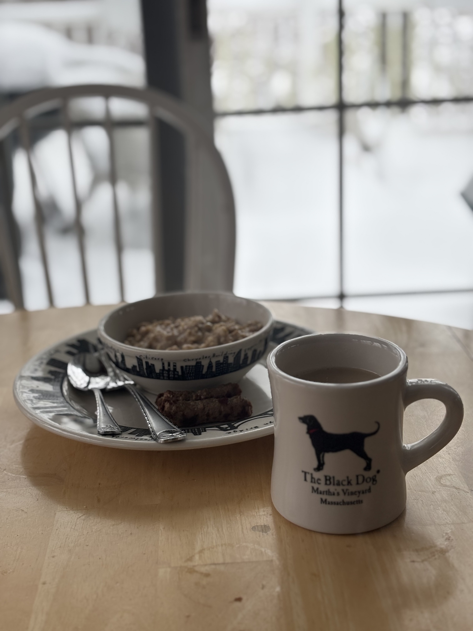 A bowl of homemade oatmeal sits on a plate with some silverware and two sausage links. In front is a Black Dog mug of coffee. In the background we look through a glass door onto a snow covered deck. 