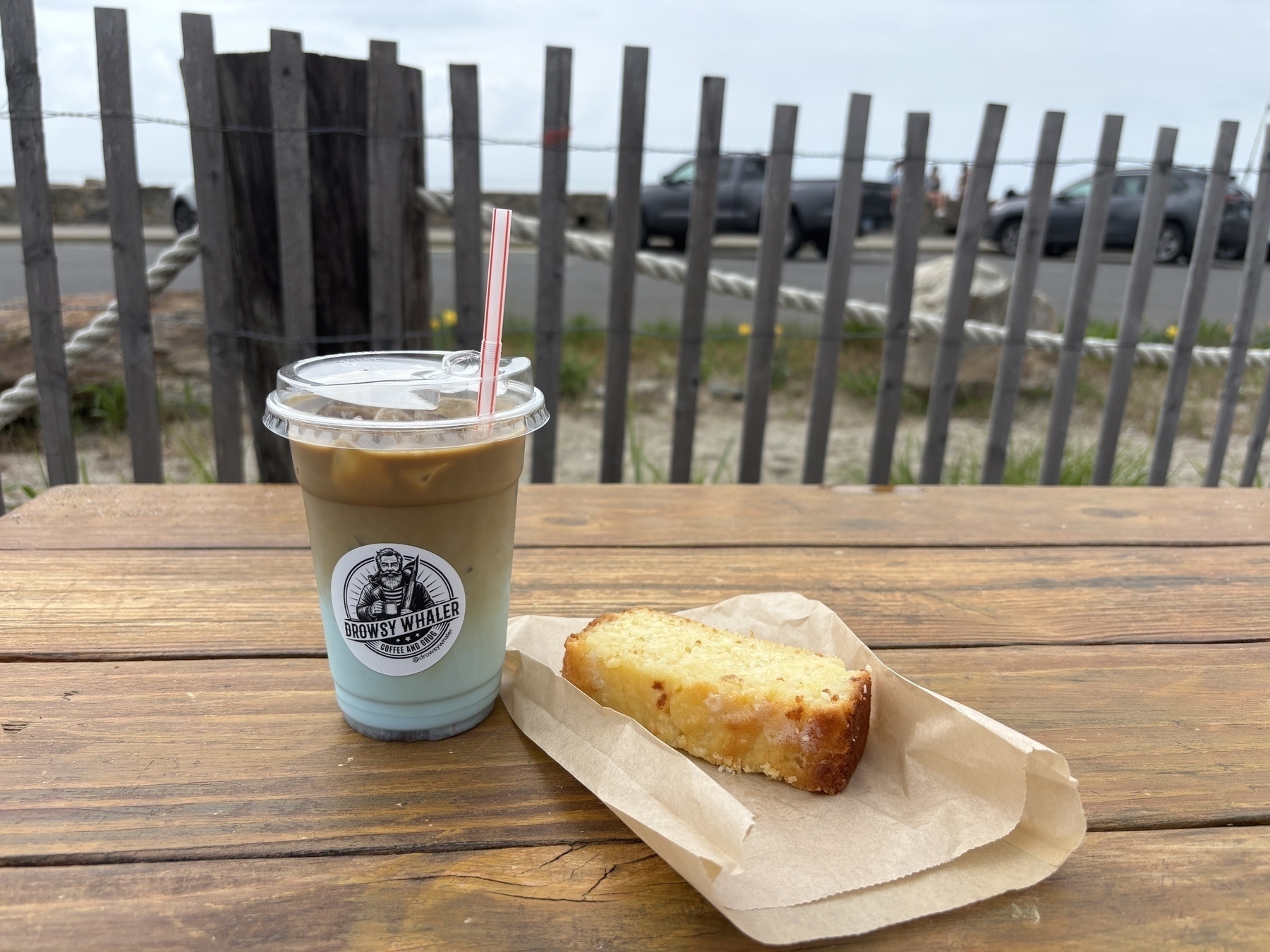The blue milk espresso sits on a picnic table with a slice of lemon pound cake to its right. A grey wooden picket fence is in the background and behind that is the sea wall and the sound. 