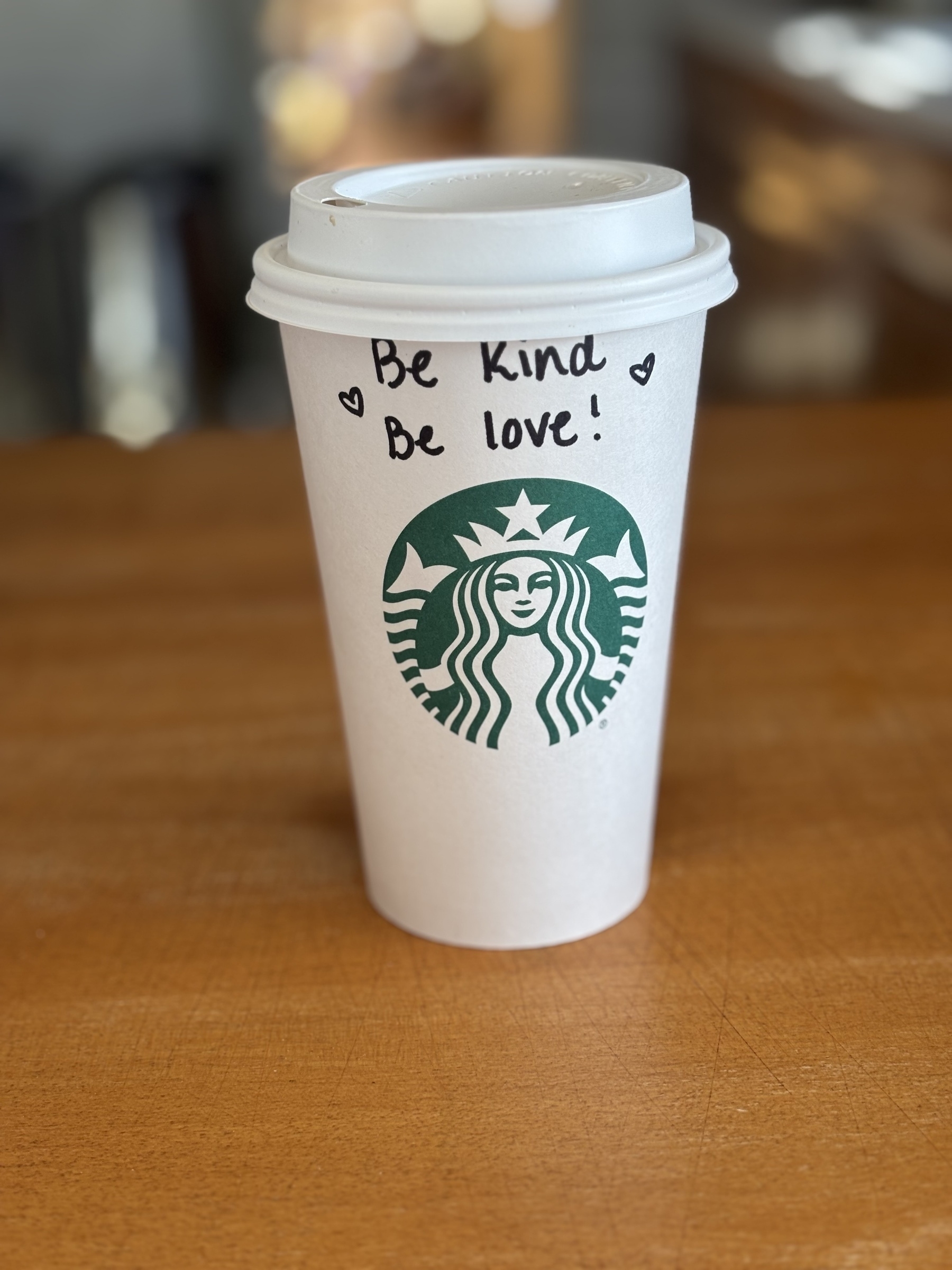 A grande white Starbucks cup with the green Starbucks logo. The barista has written “Be kind. Be love.” above the logo enclosed with hearts. 