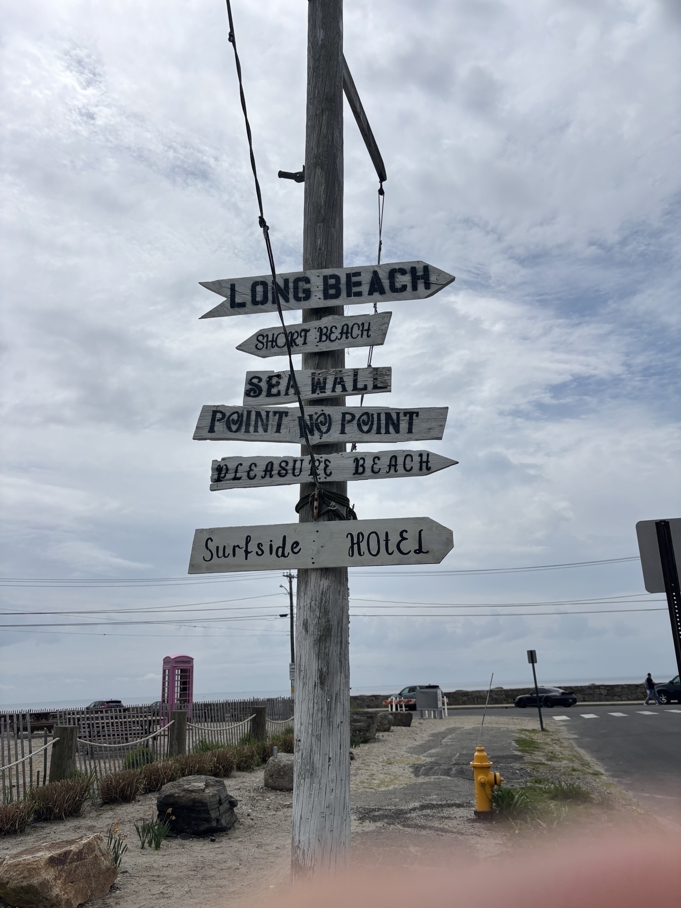 A weathered gray pole stands in front of the sea wall. There are a number of weathered gray arrow signs on it pointing in different directions. They read “Long Beach” “short beach” “sea wall” “point to point” “pleasure beach” and “surfside hotel”