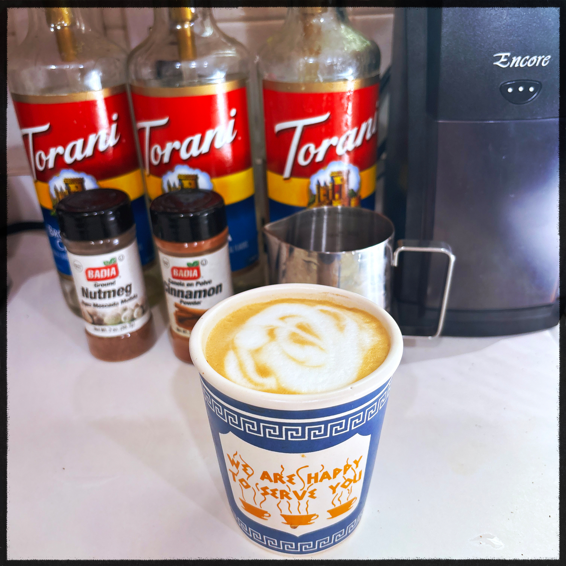 A classic “We Are Happy To Serve You” Greek diner cup (ceramic though, not paper) holds moka pot espresso with some dubious latte art. In the background are three pump bottles of Torani syrup, a metal milk pitcher and an Encore grinder. Also containers of nutmeg and cinnamon. 